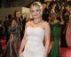 Woman in a sparkling white beaded off‑the‑shoulder gown on a red carpet, smiling at the camera, wearing a dark beaded necklace and bracelet, with guests in formal attire in the background.