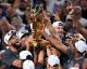 Basketball players celebrate by lifting a golden championship trophy amid cheering fans and teammates.