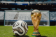 Gold FIFA World Cup trophy beside a white soccer ball on a green pitch with the goal in the background, stadium seats visible behind.
