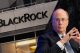 BlackRock building exterior with a large logo; a man in a dark suit speaks into a microphone at a podium in the foreground.
