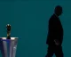 Silhouette of a man in a suit walking past a tall pedestal topped with a gleaming golden trophy against a teal background.