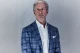 Portrait of a mature man in a blue plaid blazer and white shirt, with a white pocket square, standing against a pale background.