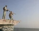 Two men with backpacks and rifles stand on the bow of a boat, aiming outward toward the sea at sunset-like light.